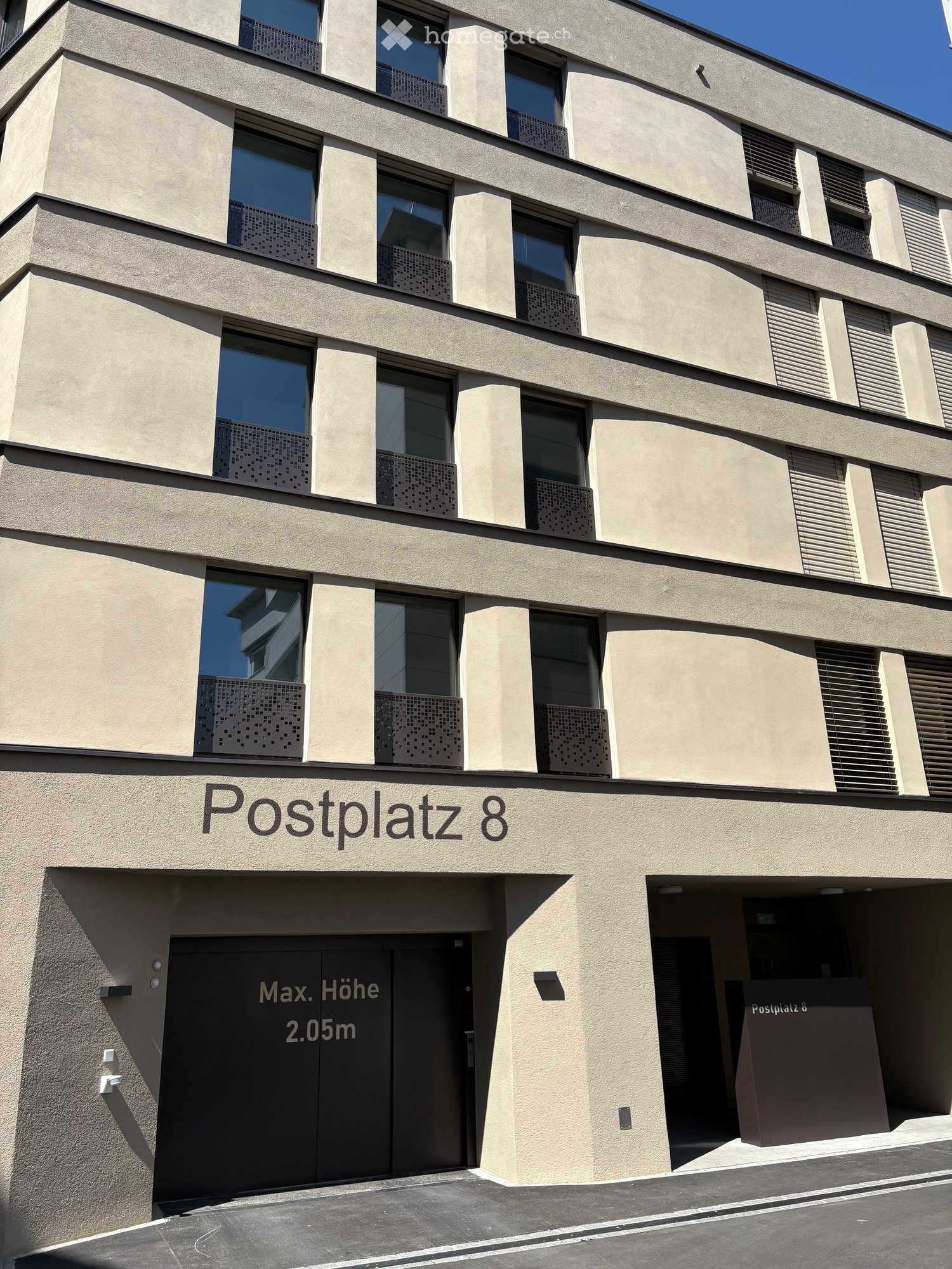 Modern multi-story building with beige facade, large windows, and decorative metal grilles. Sign reads "Postplatz 8" and indicates maximum height of 2.05m.