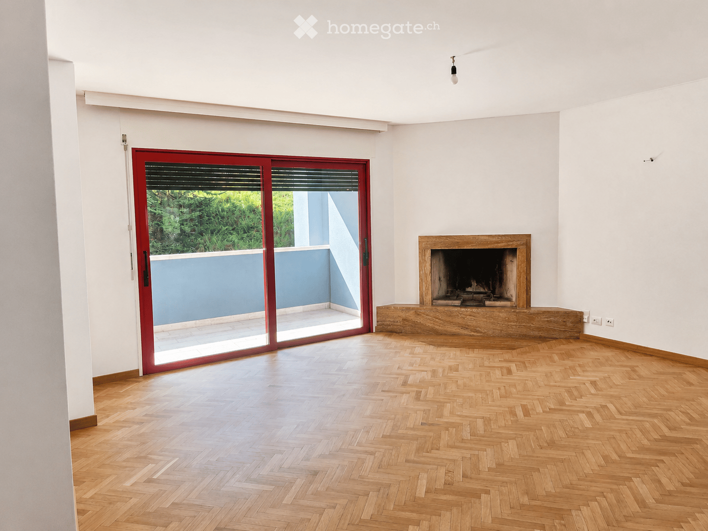 Empty room with wooden herringbone flooring, white walls, a fireplace, and a large red sliding glass door leading to a balcony or outdoor area.