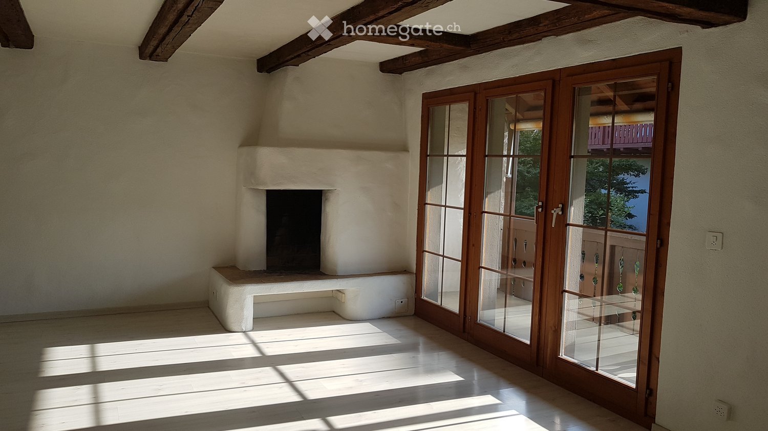 Empty room with wooden floor, exposed wooden beams, fireplace, and large glass doors leading to a balcony.