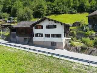 A traditional Swiss-style building with a pitched roof and wooden facade, situated in a green, mountainous area with a stone pathway leading to it.