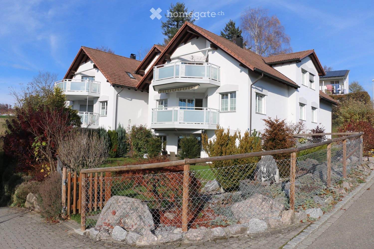 A two-story white house with a red-tiled roof, multiple balconies, and a fenced garden area with shrubs and trees.