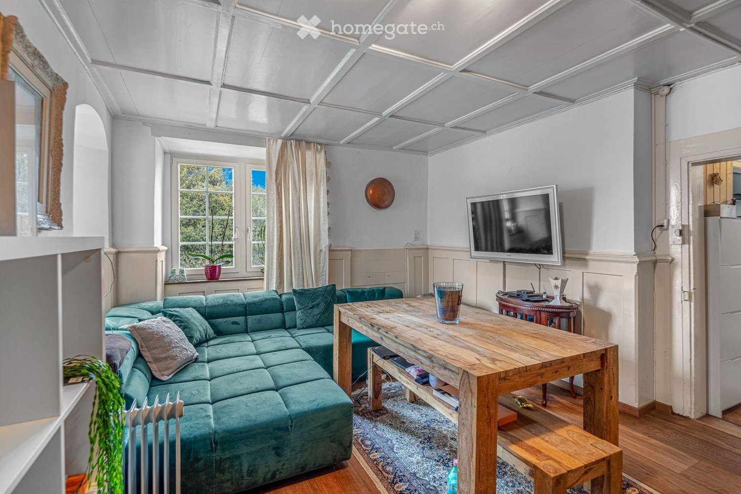 Bright living room with a green sectional sofa, wooden coffee table, flat-screen TV, side table, and decorative items.