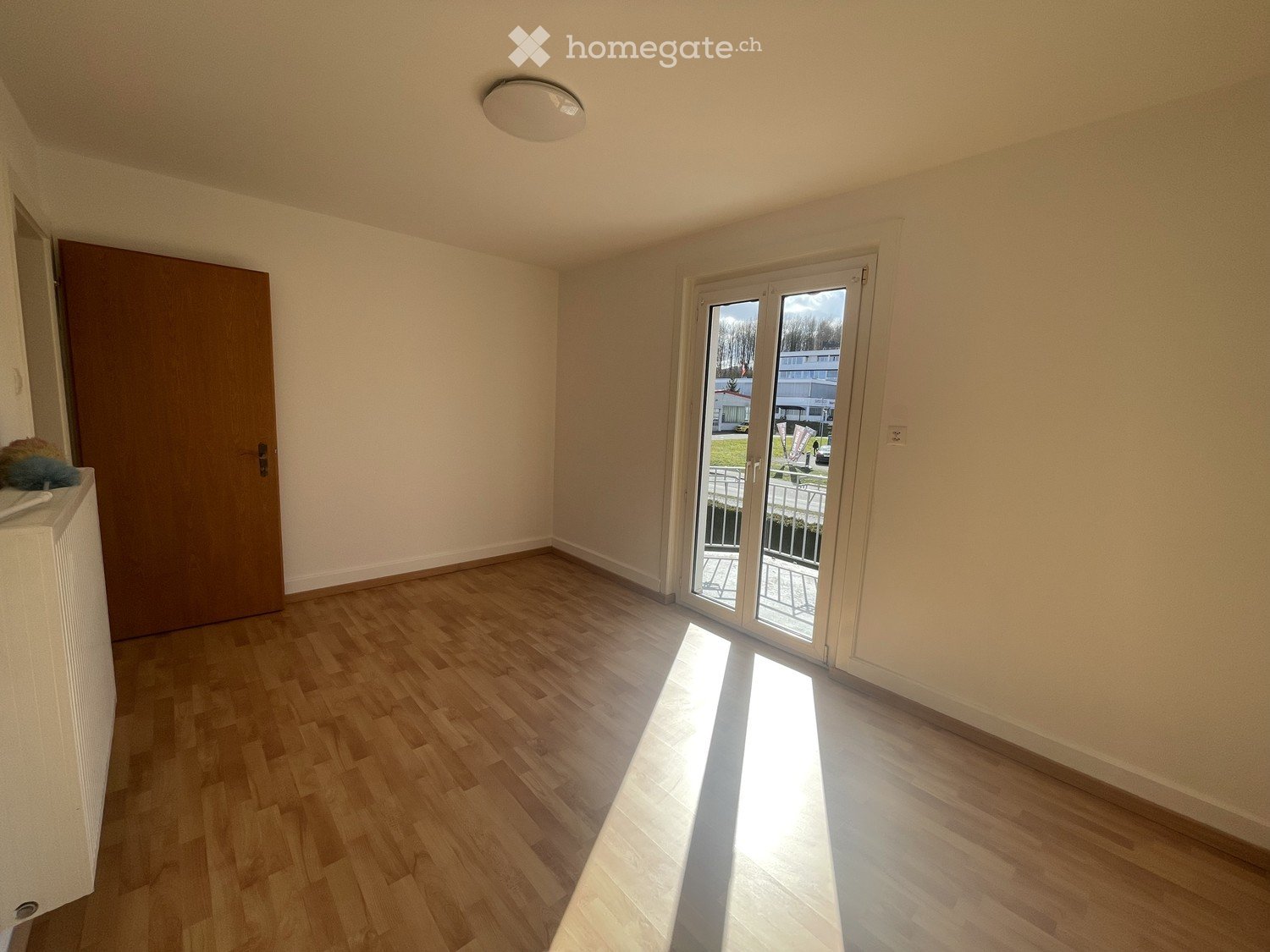 Empty room with wooden flooring, white walls, a sliding glass door leading to a balcony, and a closed wooden door.