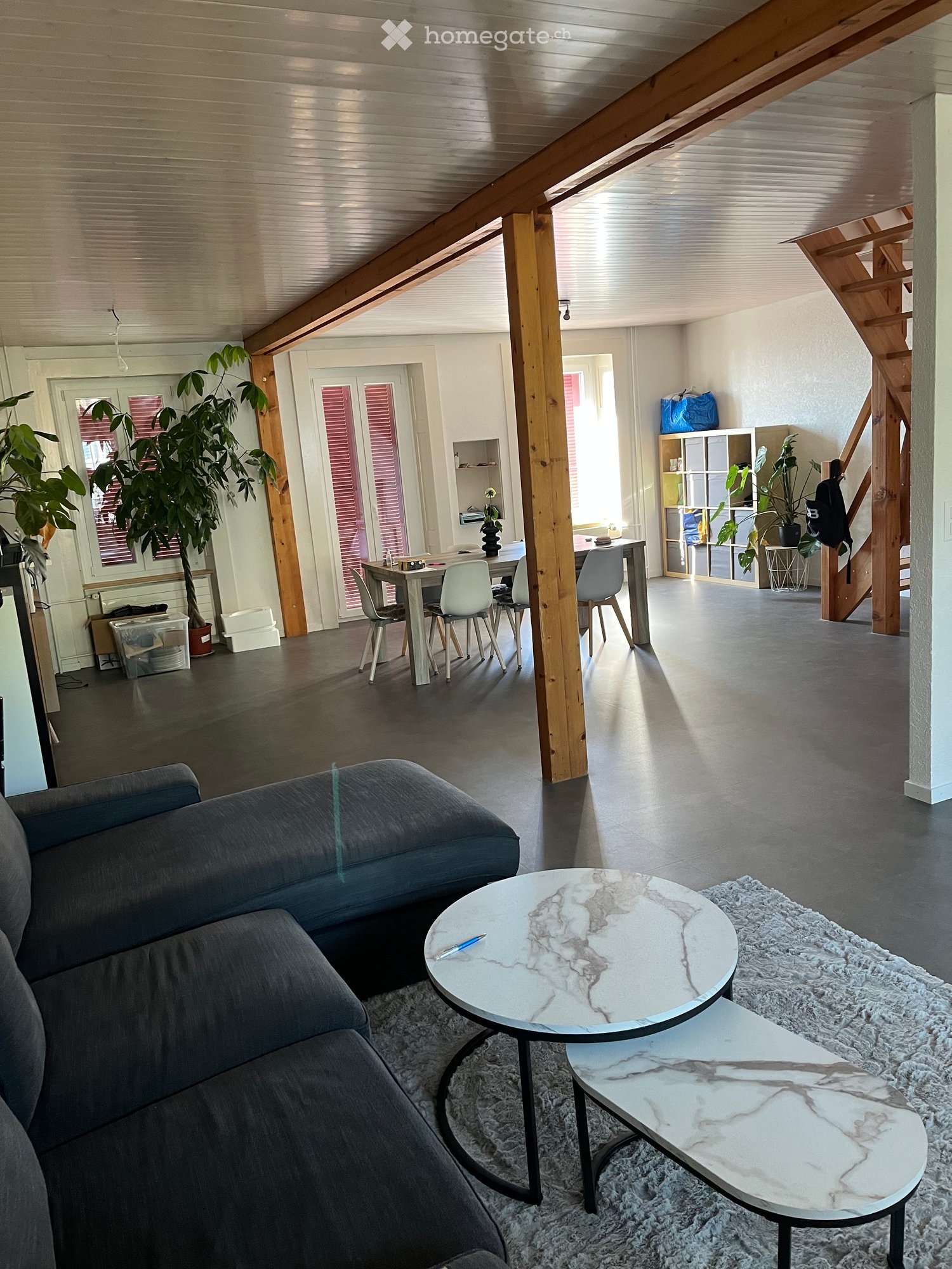 Open-plan living and dining area with exposed wooden beams, grey sofa, marble coffee table, dining table with chairs, and shelving unit with plants.