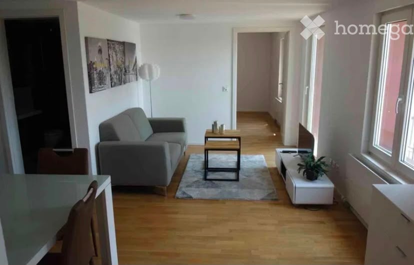 Modern living room with light wooden flooring, grey sofa, coffee table, small side table, and a plant on a white console. The room features large windows and a hallway leading to another area.