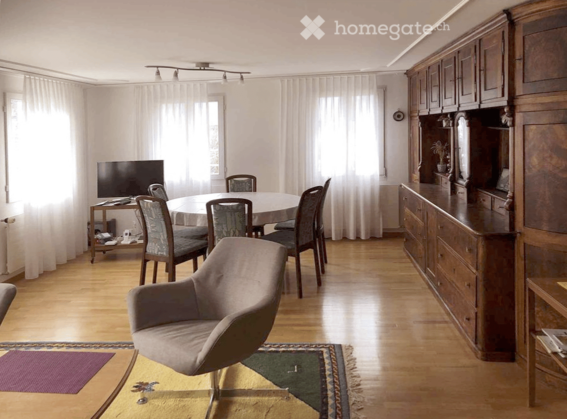 The image shows a spacious open-plan living and dining area. The room features a large wooden cabinet/entertainment unit along one wall, a round dining table with chairs, and a comfortable gray sofa. The floors are hardwood, and there are large windows pr
