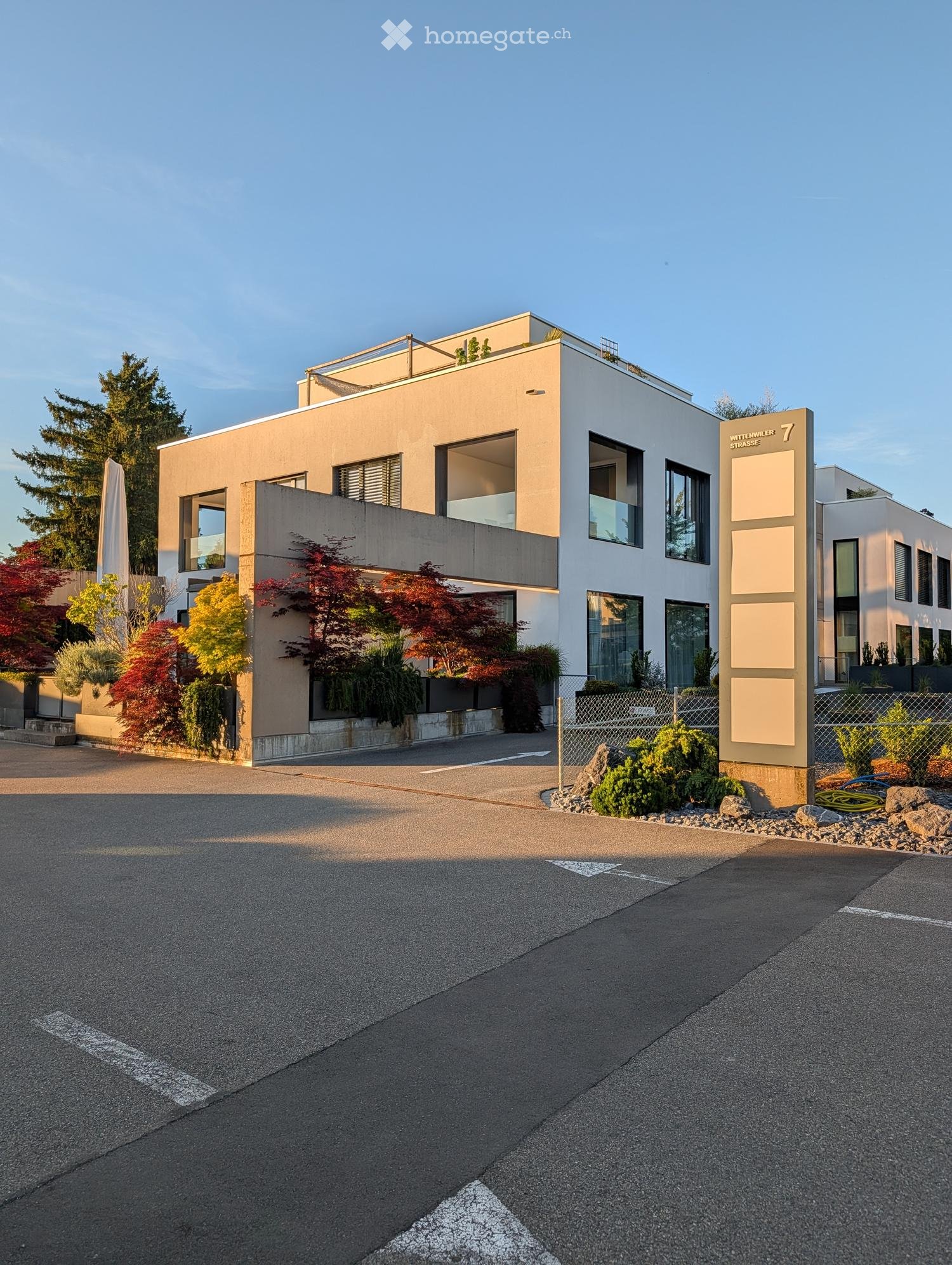 Modern two-story building with a combination of beige and white exterior, large windows, and a balcony on the upper level. The building is surrounded by landscaped greenery and a paved driveway.