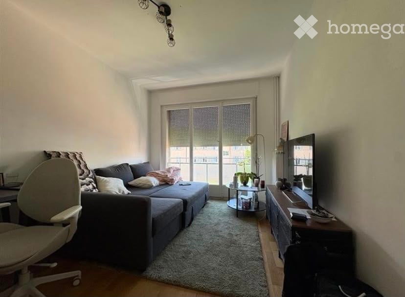 Modern living room with a grey sectional sofa, coffee table, TV stand, floor lamp, and a small round table near the balcony. The room features large windows with blinds and a cozy rug on the floor.