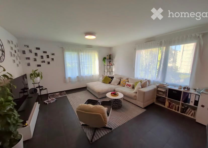 Modern living room with a large sectional sofa, coffee table, armchair, bookshelf, and decorative plants. The room features large windows with white curtains and a dark wooden floor.