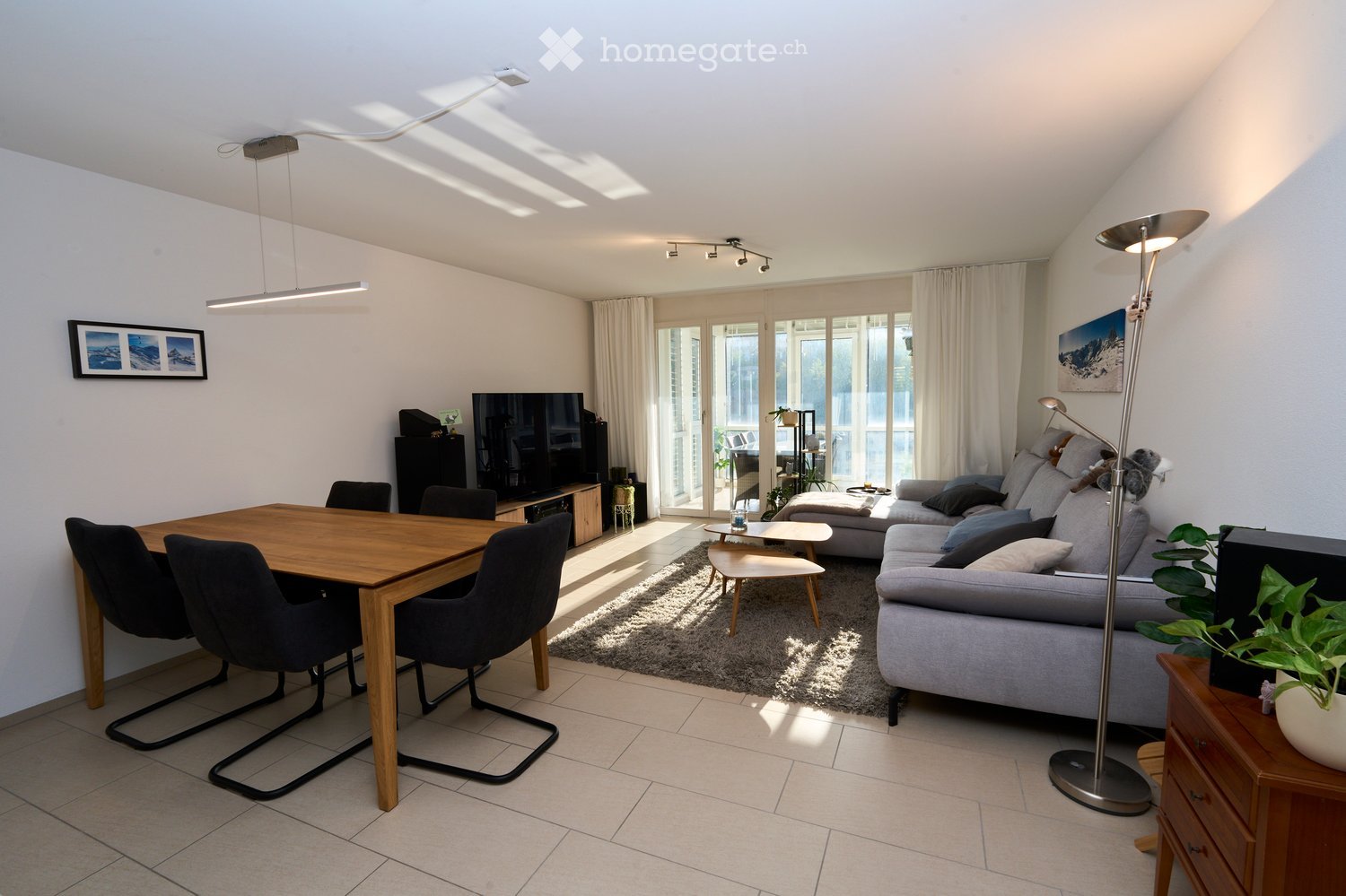 Bright living room with a grey sectional sofa, wooden dining table with four chairs, a coffee table, a television, and a floor lamp. Large windows and sliding glass doors lead to a balcony.