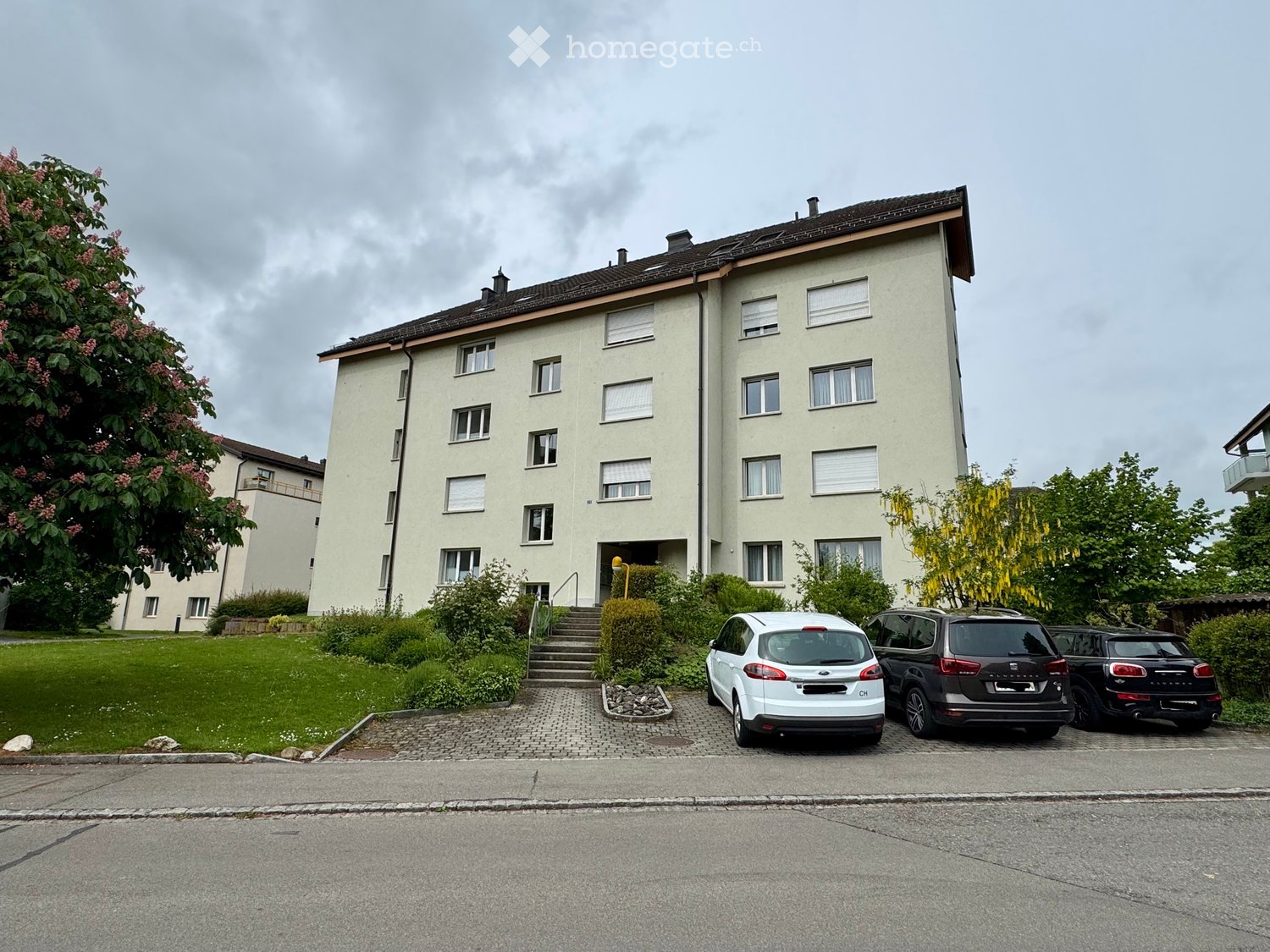 Four-story residential building with multiple windows, a staircase leading to the entrance, and a small garden area with shrubs and trees. Three cars are parked in front of the building.