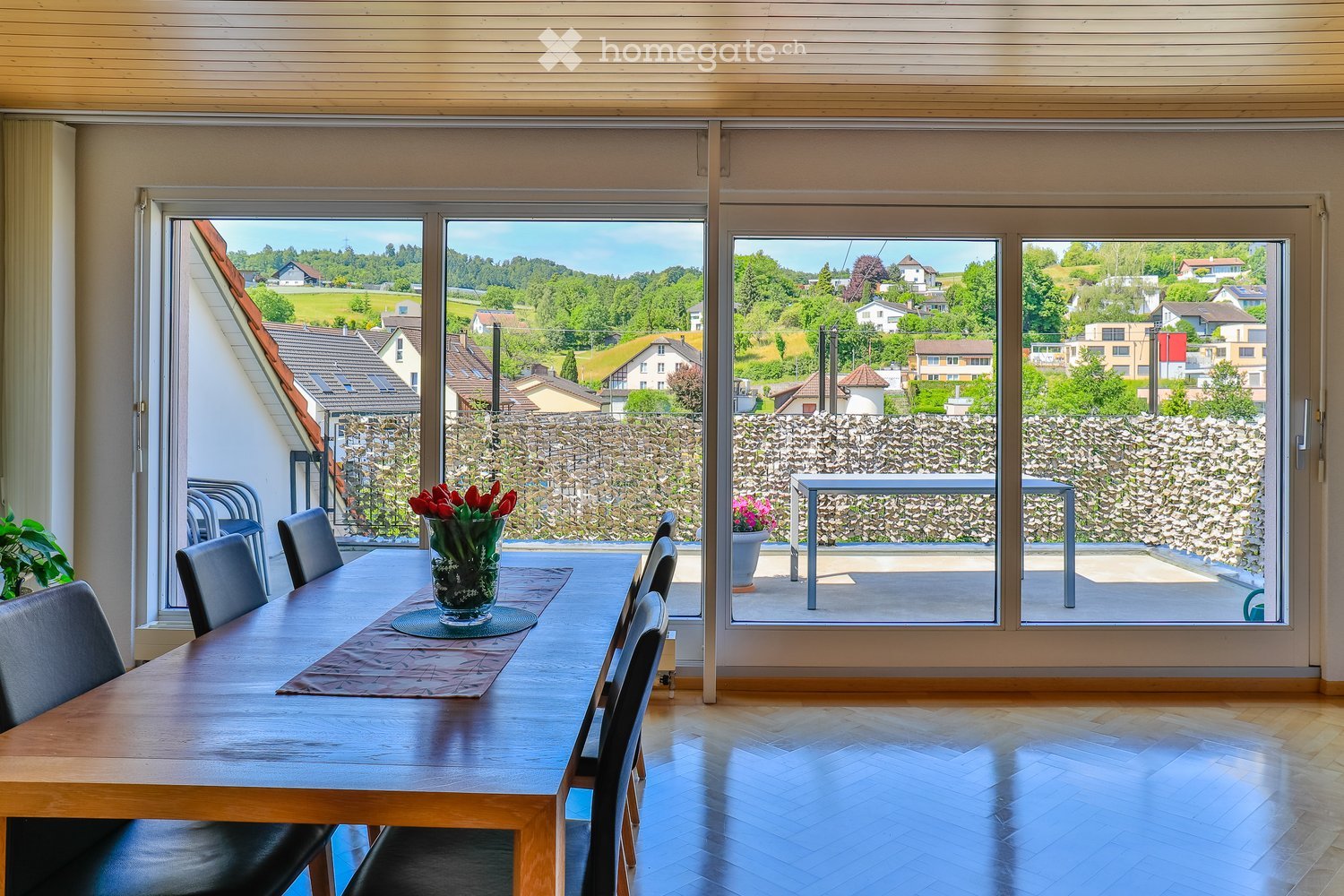 Bright dining area with a wooden table, black chairs, and a vase of red tulips, overlooking a scenic countryside view through large windows.