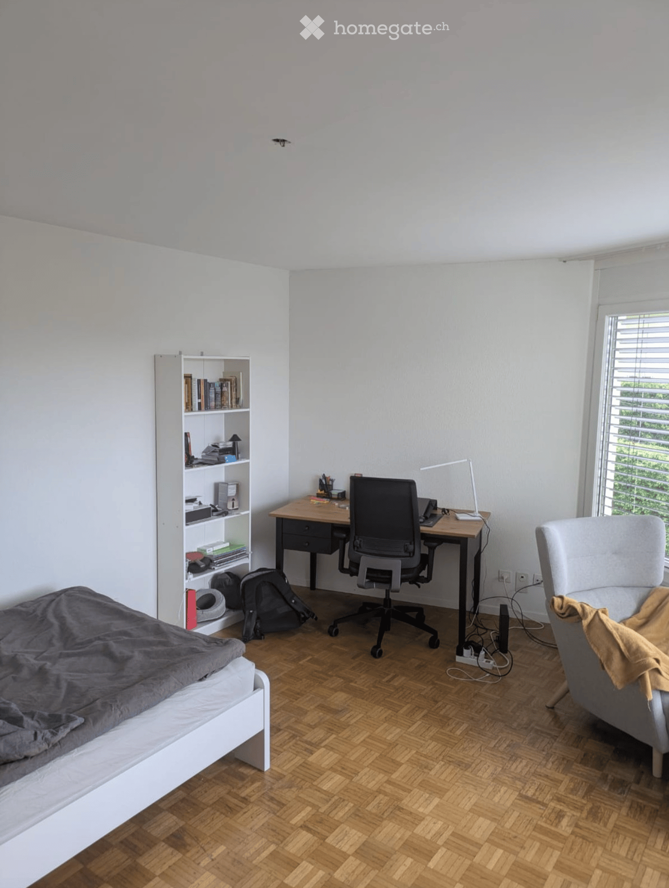 A modern, multi-functional room with a bed, desk, and seating area. Features include a wooden floor, white walls, a large window with blinds, and a bookshelf with books and storage items.