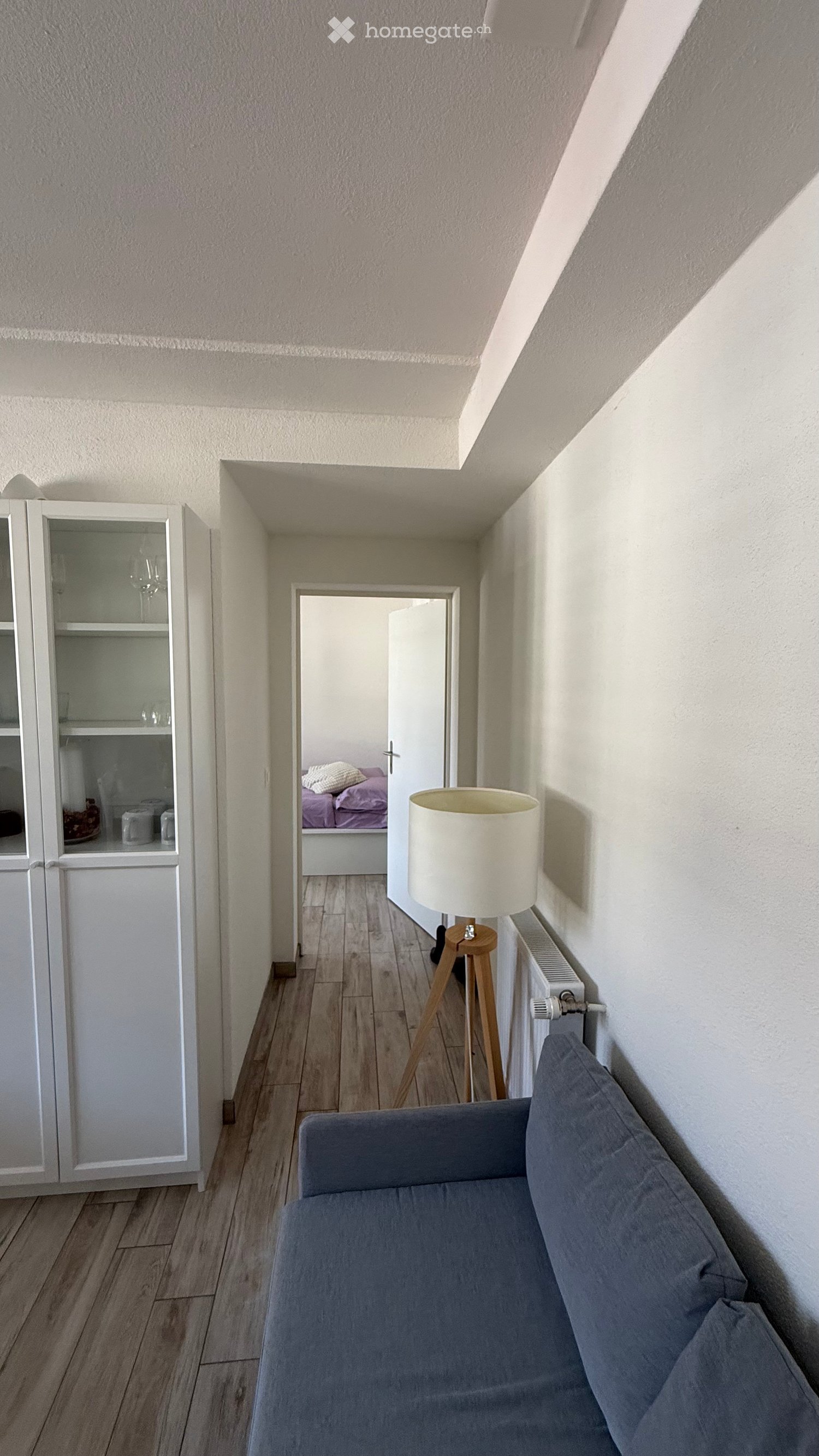 A small, modern living space with a grey sofa, a wooden table lamp, a white cabinet, and a radiator. The room has wooden flooring and leads to a bedroom with purple bedding.