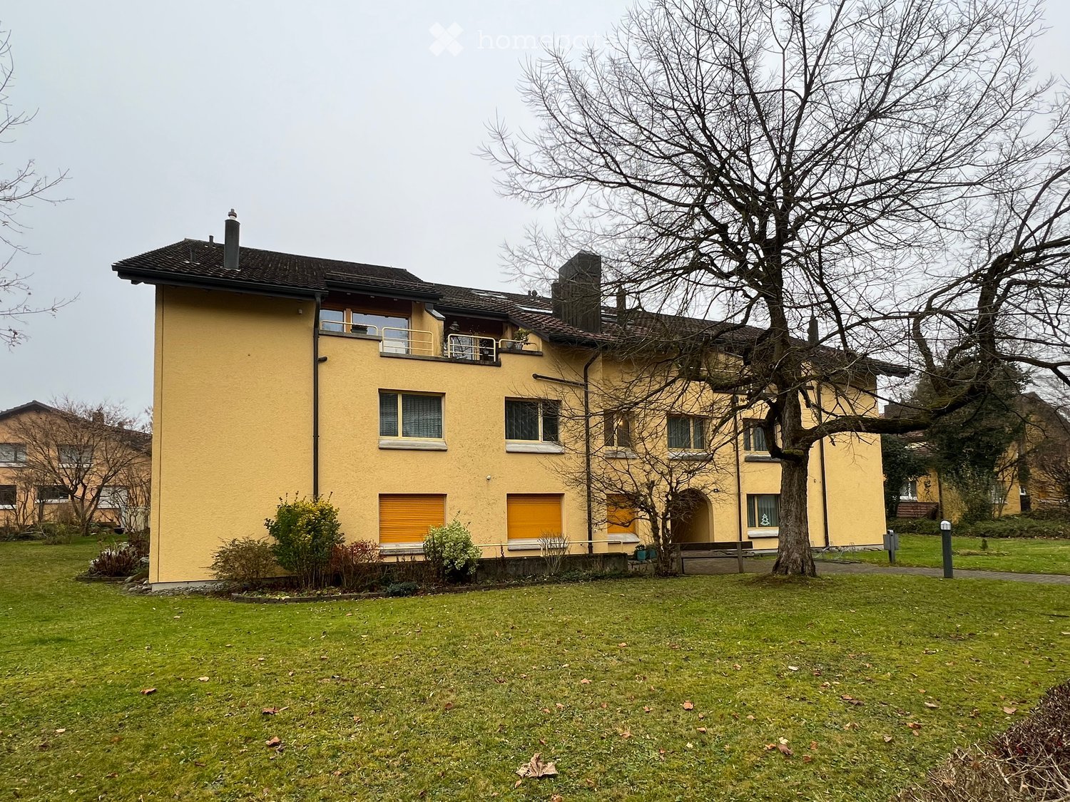 Two-story yellow house with multiple windows, a balcony, and a chimney, surrounded by a grassy lawn and trees.
