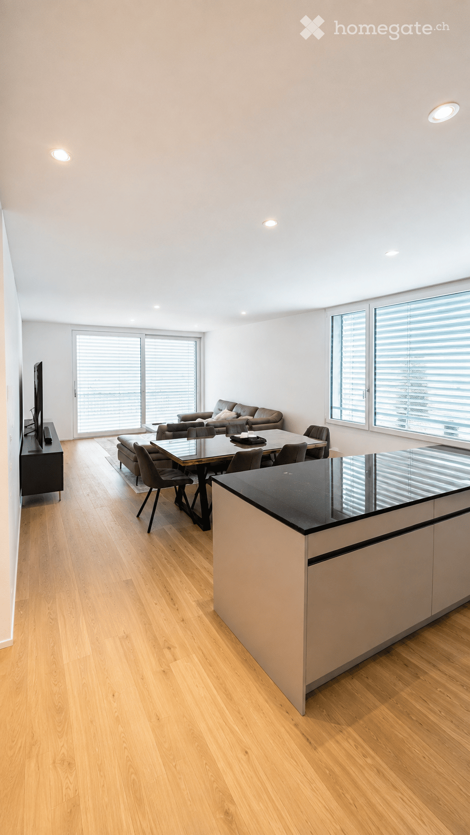 Open-plan living and dining area with light wooden flooring, large windows, a sofa, dining table with chairs, and a modern kitchen island.