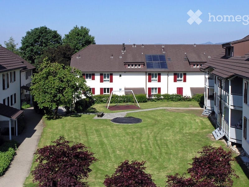 Well-maintained lawn with trimmed hedges and trees, surrounded by residential buildings with red shutters and solar panels on the roof.