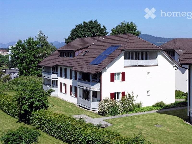 A modern two-story house with white walls, red shutters, and a brown tiled roof. Features include multiple balconies, a solar panel on the roof, and a well-maintained lawn with bushes and trees.