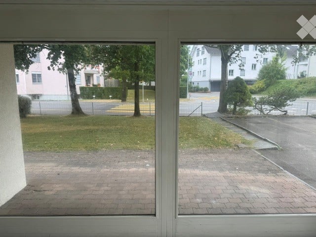 Empty room with large windows showing a view of a paved area, trees, and neighboring buildings.
