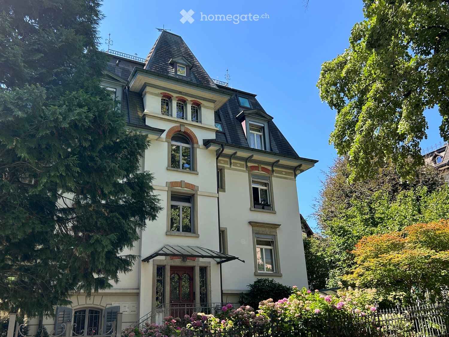 A three-story residential house with a pitched roof, multiple windows, and decorative architectural details. The house is surrounded by lush greenery and flowering bushes.