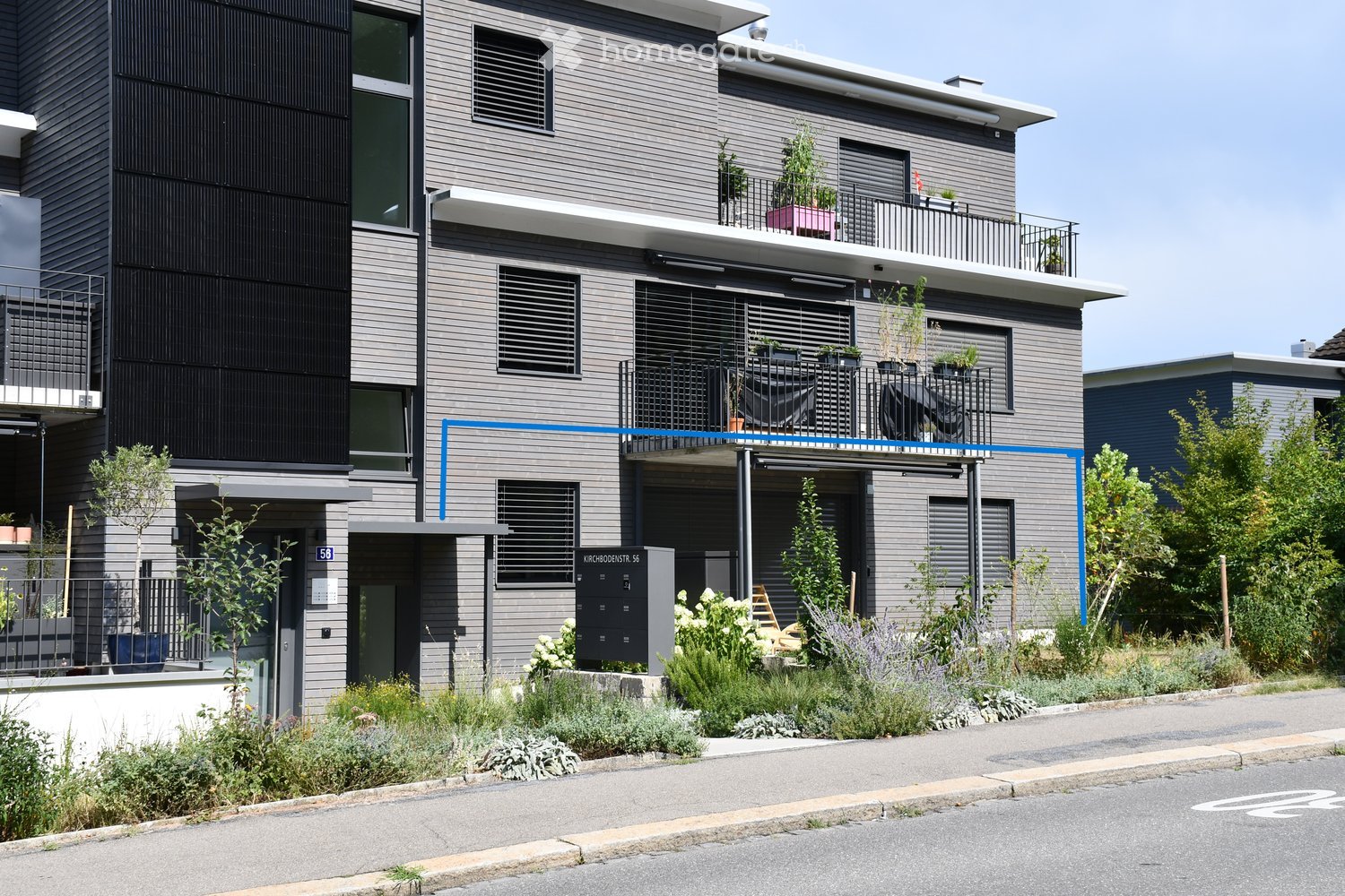Modern two-story building with grey siding, multiple balconies, and greenery in front.