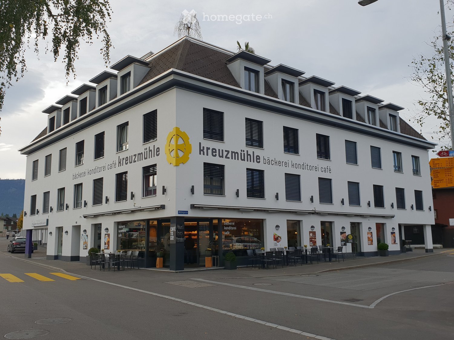 Three-story building with a modern facade, large windows, and a sign reading "Kreuzmühle Bäckerei Konditorei Café". The ground floor features a café with outdoor seating, tables, and chairs.