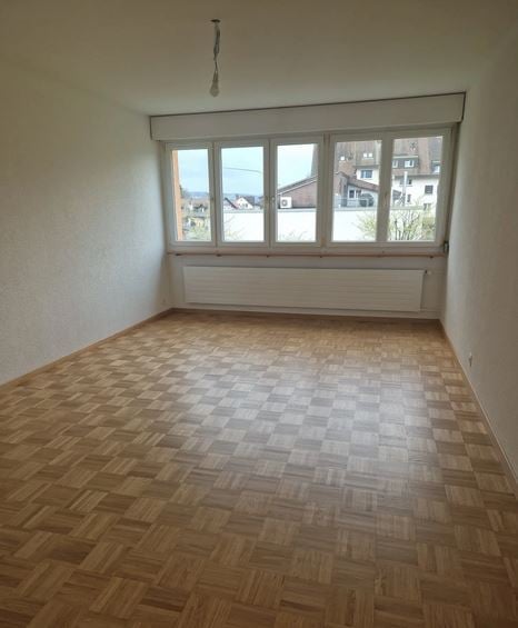 Empty room with wooden parquet flooring, white walls, large windows, and a radiator below the windows.