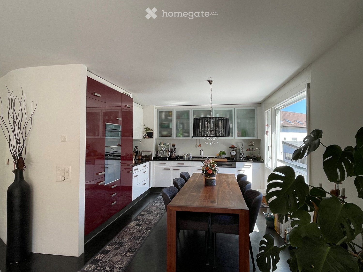 Modern kitchen with red and white cabinets, a wooden dining table with chairs, a chandelier, and decorative plants.