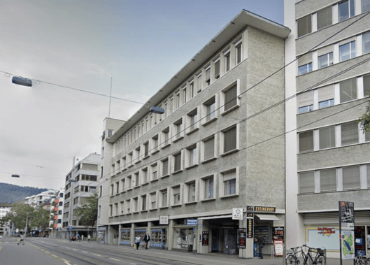 Multi-story building with a flat roof and numerous windows, located on a street with a tram line running alongside.