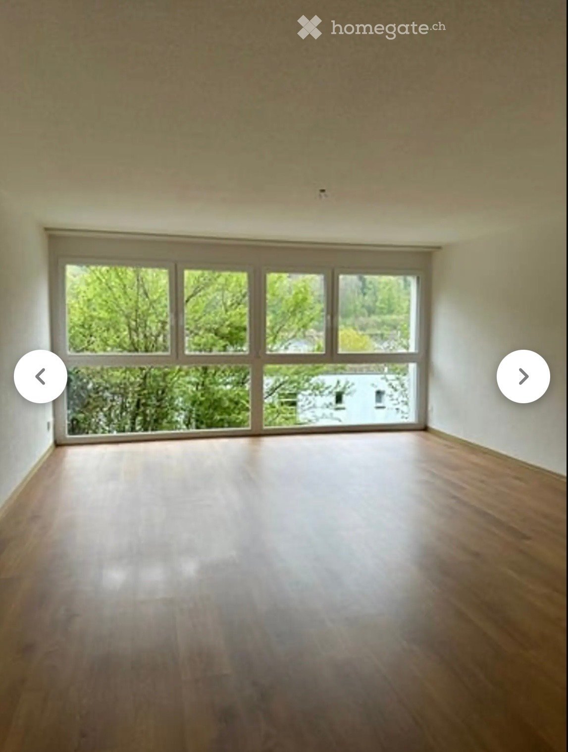 Empty room with wooden flooring and large windows showing greenery outside.