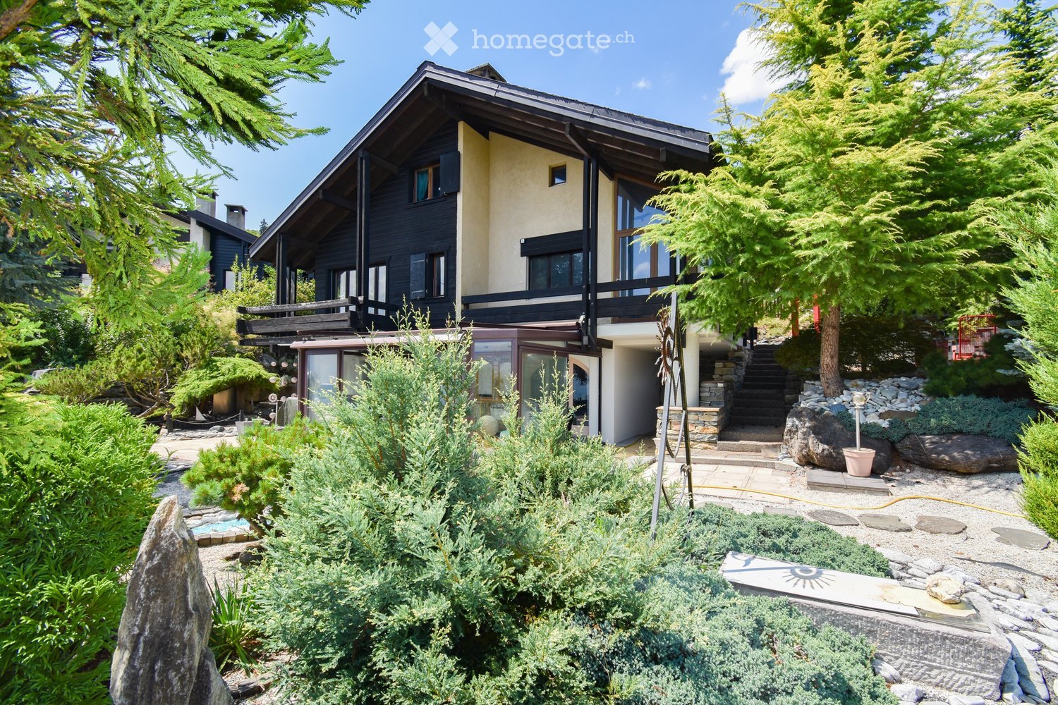 A two-story house with a modern design, featuring a combination of dark wood and light-colored walls, large windows, and a balcony on the upper level. The house is surrounded by well-maintained gardens with various shrubs, trees, and a stone pathway leadi