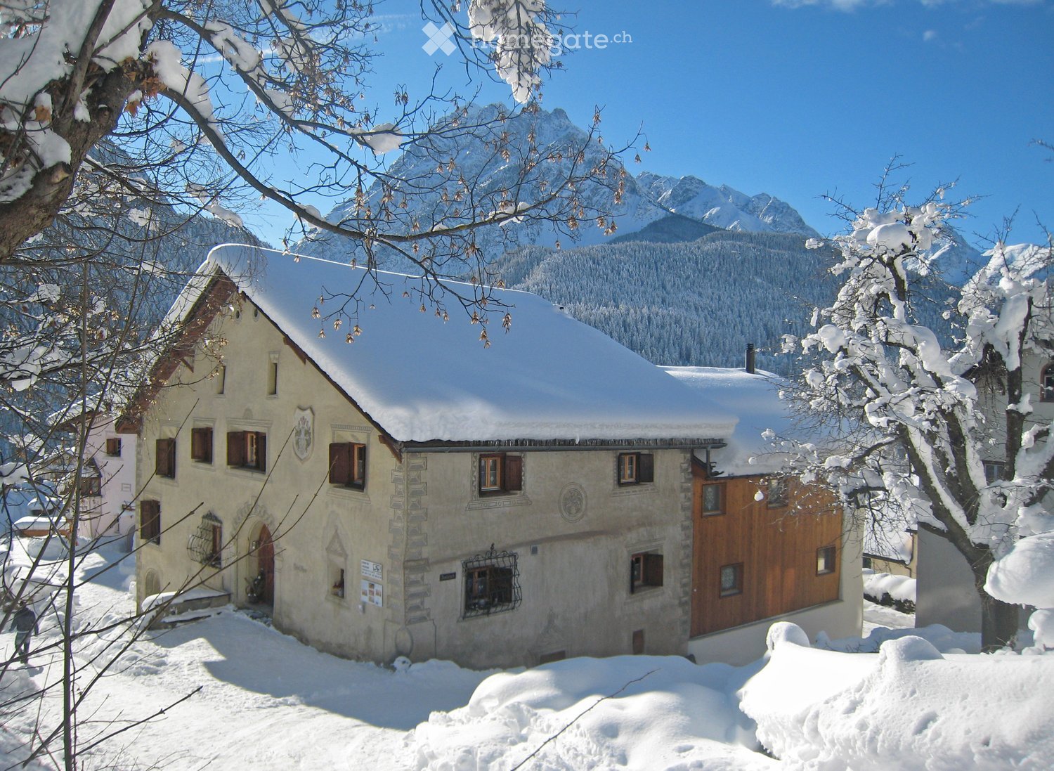 A two-story house with a snow-covered roof and wooden shutters, surrounded by snow-covered trees and mountains.