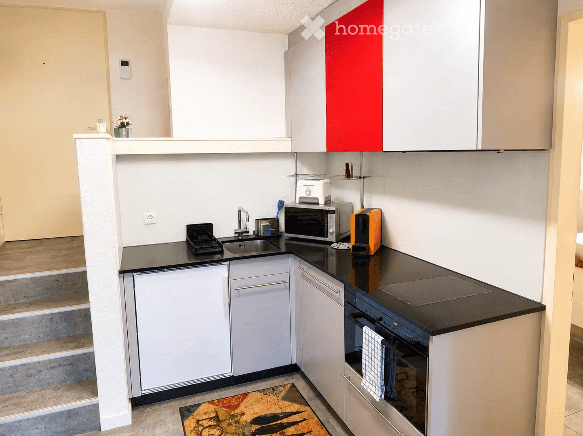 Modern kitchen with white cabinets, black countertops, and stainless steel appliances including a microwave, oven, and toaster. A small sink with a faucet is visible, along with a colorful rug on the floor.