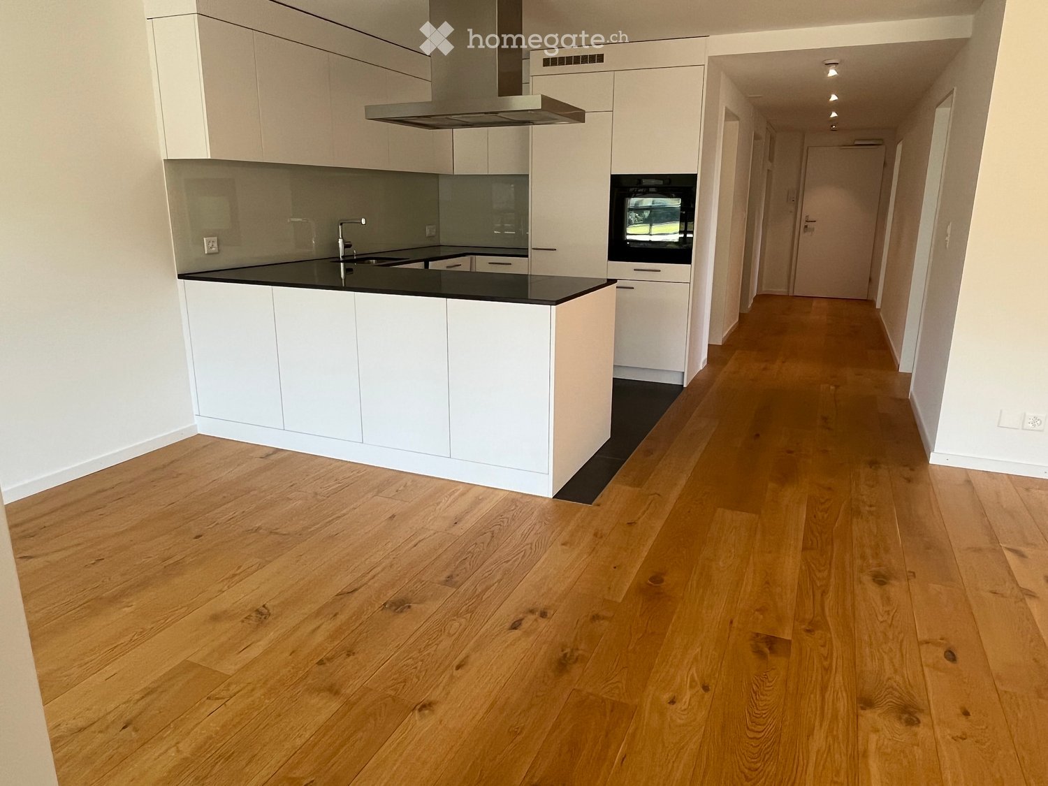 Empty hallway with wooden flooring and white walls, leading to a modern kitchen area with white cabinets, a black countertop, and stainless steel appliances.