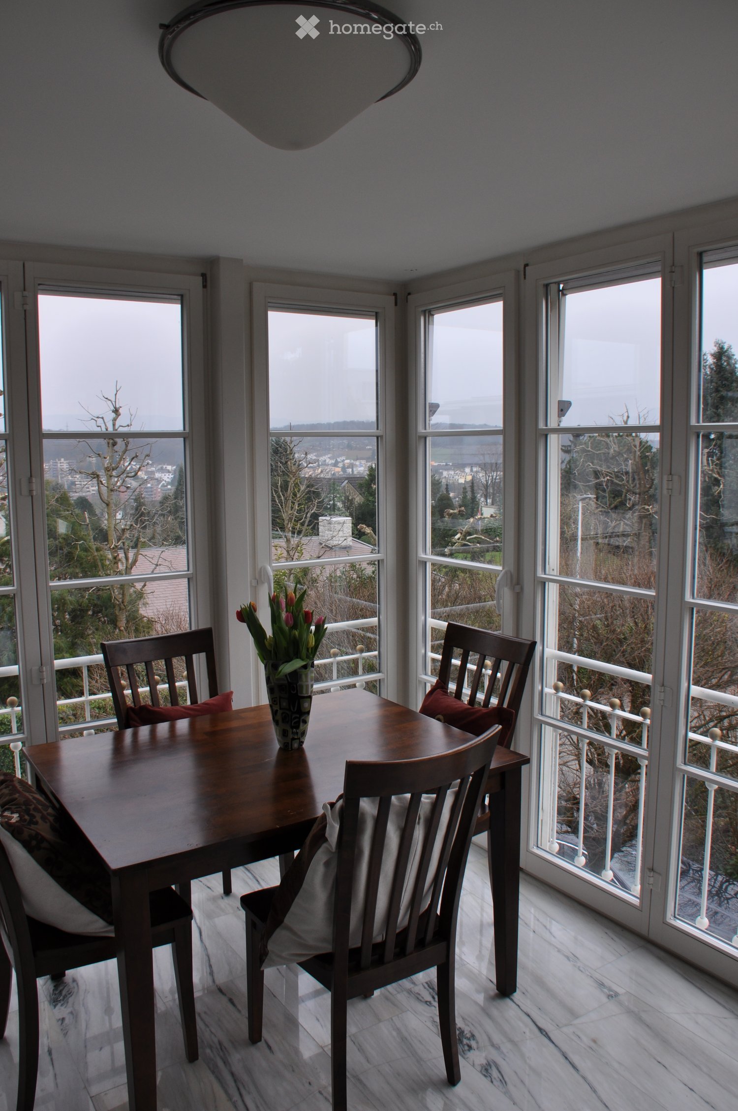 Bright dining area with large windows offering a scenic view, featuring a wooden dining table, chairs, and a vase with flowers.