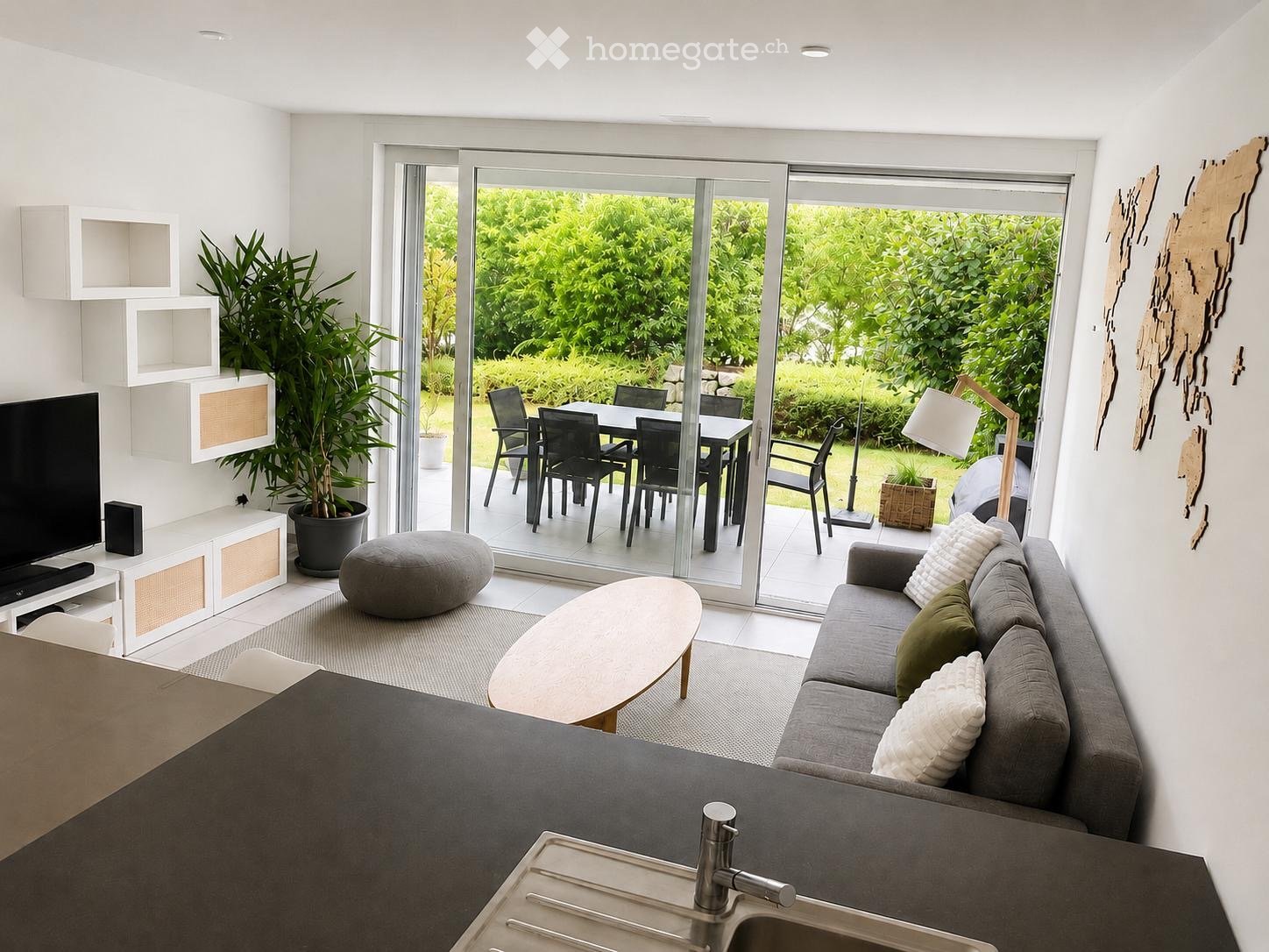 Modern living room with a grey sofa, coffee table, pouffe, TV stand, and a kitchen counter with a sink. Large glass sliding doors open to a dining area with a table and chairs.