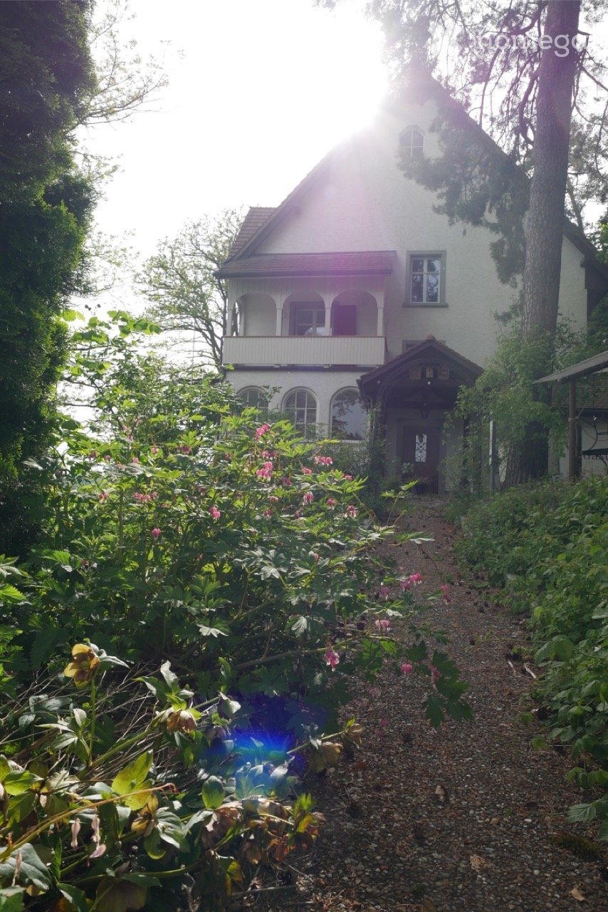 A two-story white house with a balcony and arched windows, surrounded by lush greenery and a gravel pathway.