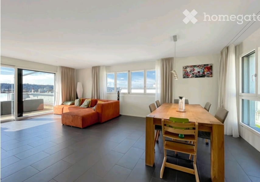 Open-plan living and dining area with large windows offering a view, orange sectional sofa, ottoman, wooden dining table with chairs, and a floor lamp.