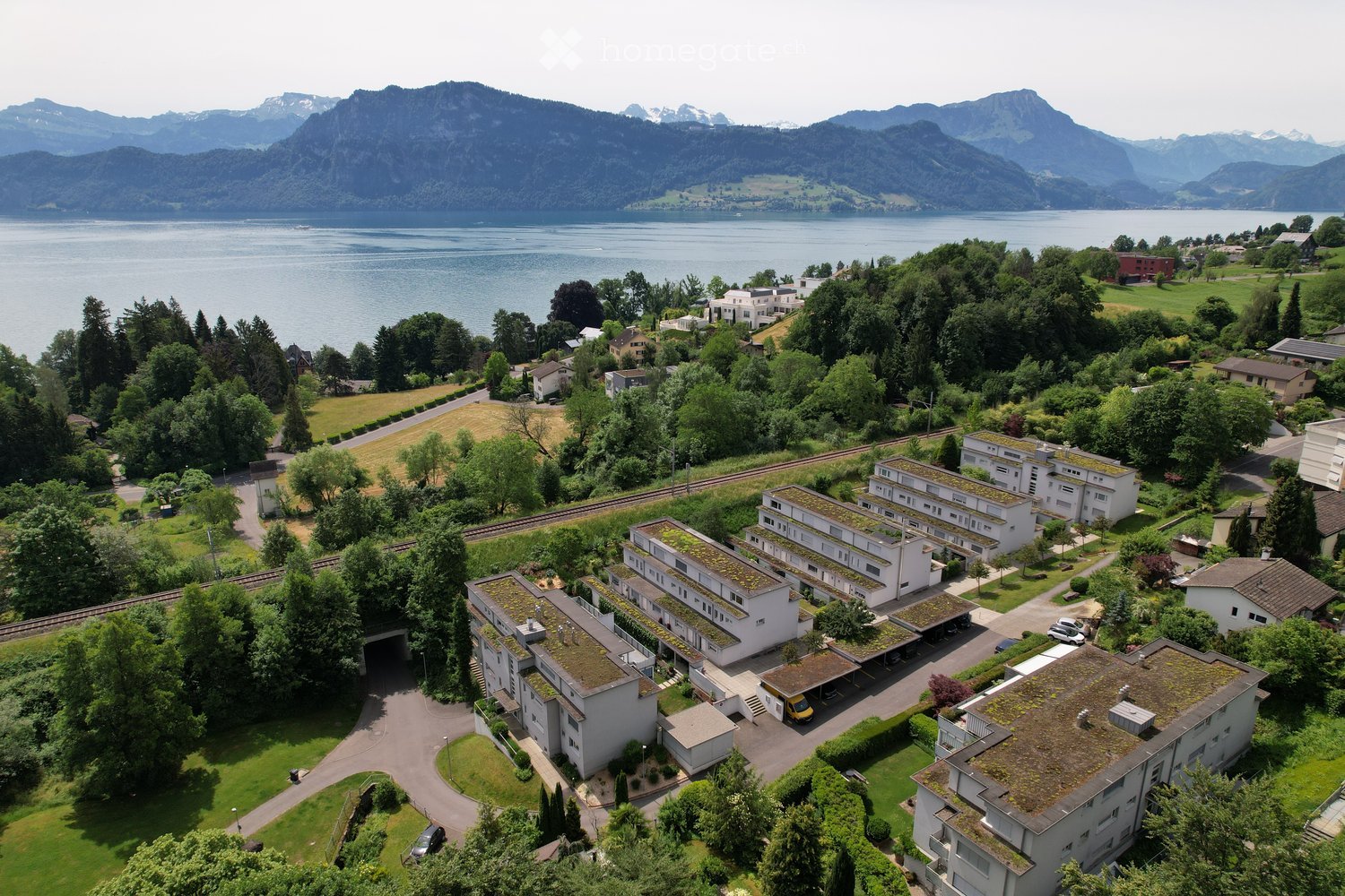 Aerial view of a residential complex with multiple buildings, surrounded by greenery, a parking area, and a scenic lake and mountain backdrop.