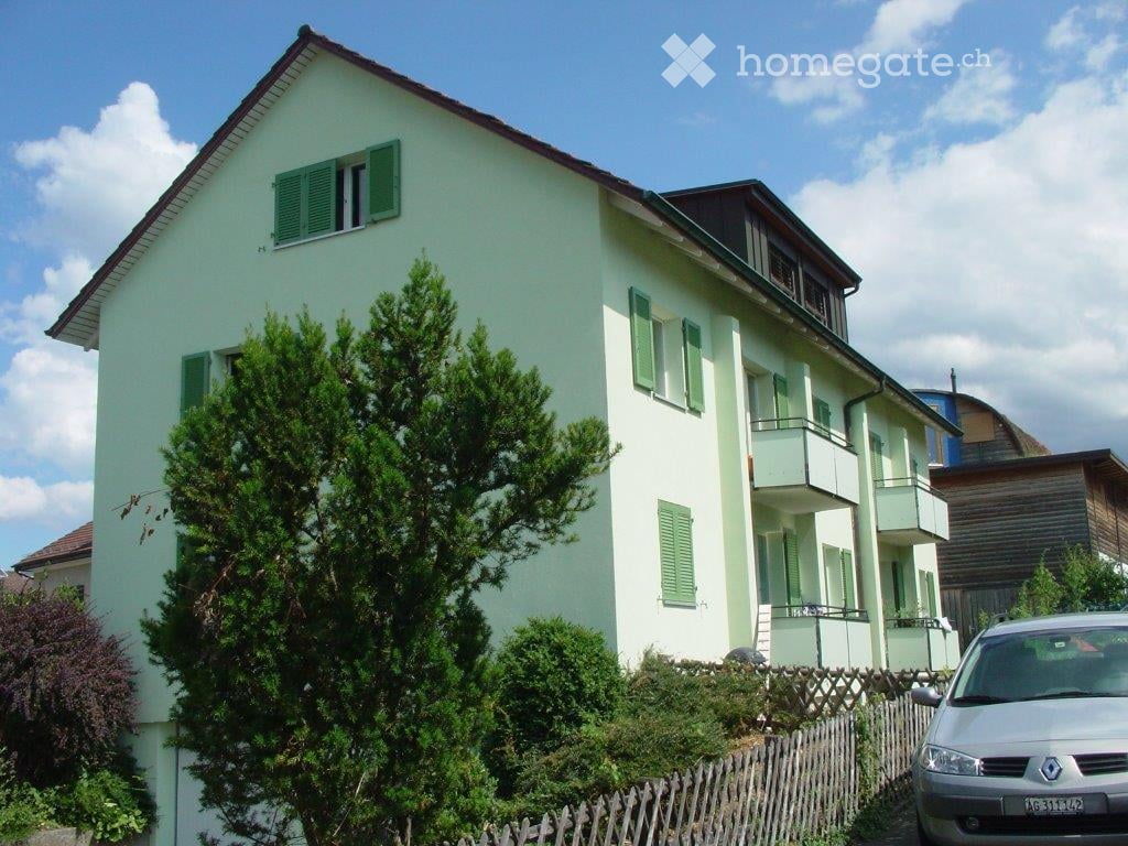 Two-story house with light green exterior, green shutters, and a balcony on the upper floor. A small tree and shrubs are in the front yard, and a car is parked nearby.