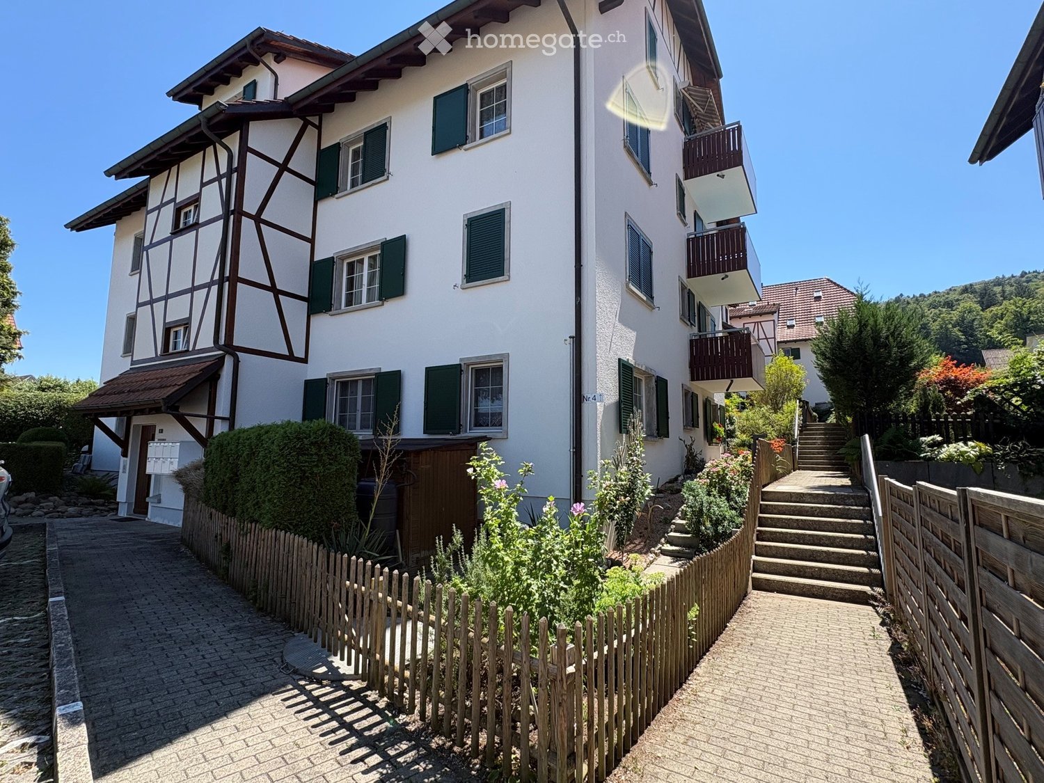 Two-story house with white walls, green shutters, and a wooden balcony. The house features a mix of traditional and modern architectural elements, with a well-maintained garden and a paved pathway leading to the entrance.