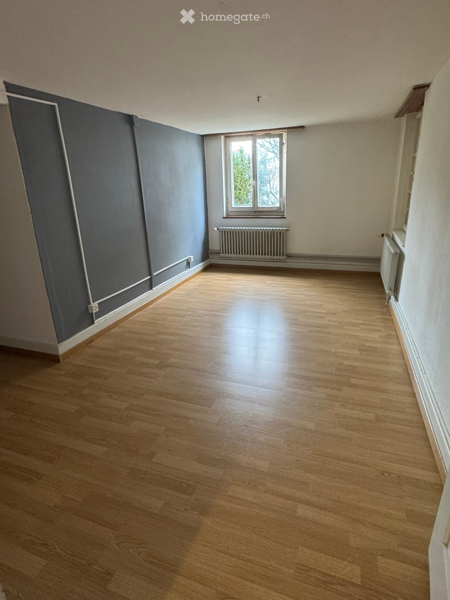 Empty room with wooden flooring, grey walls, and a single window with radiator below it.