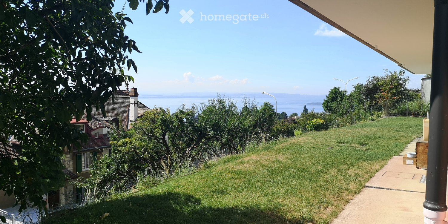 Lush garden with a pathway leading to a scenic view of a lake and distant hills, surrounded by trees and greenery.