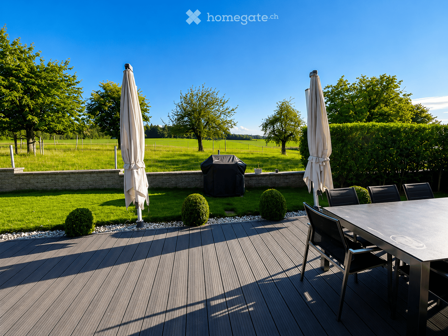 A wooden deck with a dining table, chairs, and umbrellas, surrounded by a well-maintained lawn, trees, and a stone wall.
