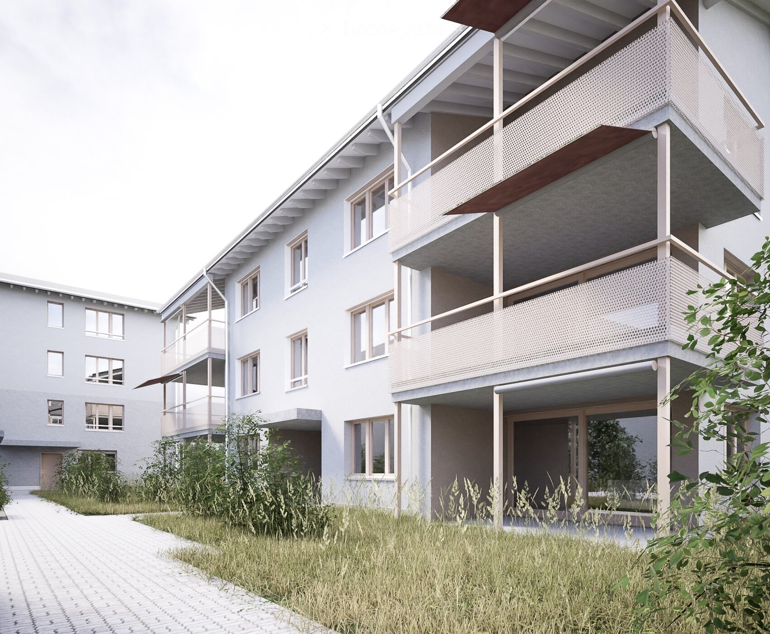 Modern two-story residential building with light gray facade, multiple windows, and balconies with perforated metal railings. The ground floor has a covered entrance area, and the surrounding area includes a paved walkway and some greenery.
