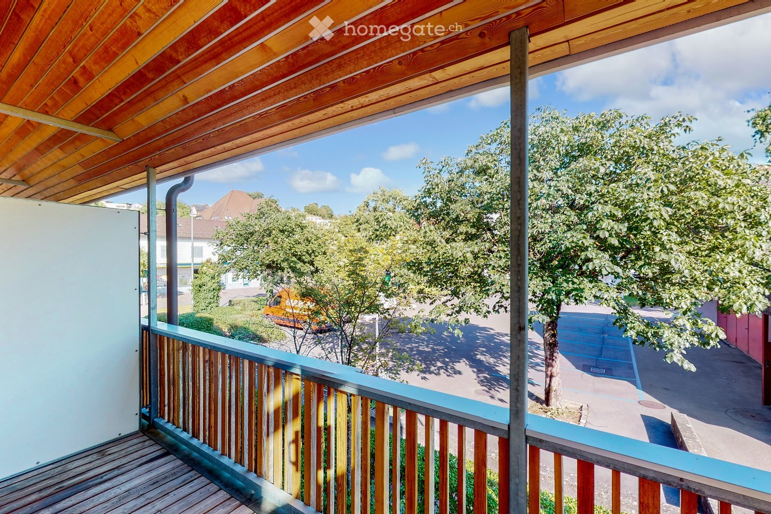 Balcony with wooden decking, railing, and a view of a tree-lined street and neighboring houses.