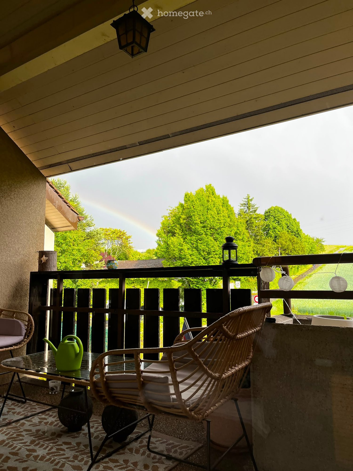 Balcony with wicker chairs, glass table, green teapot, and hanging lanterns, overlooking a green landscape with trees and a rainbow.