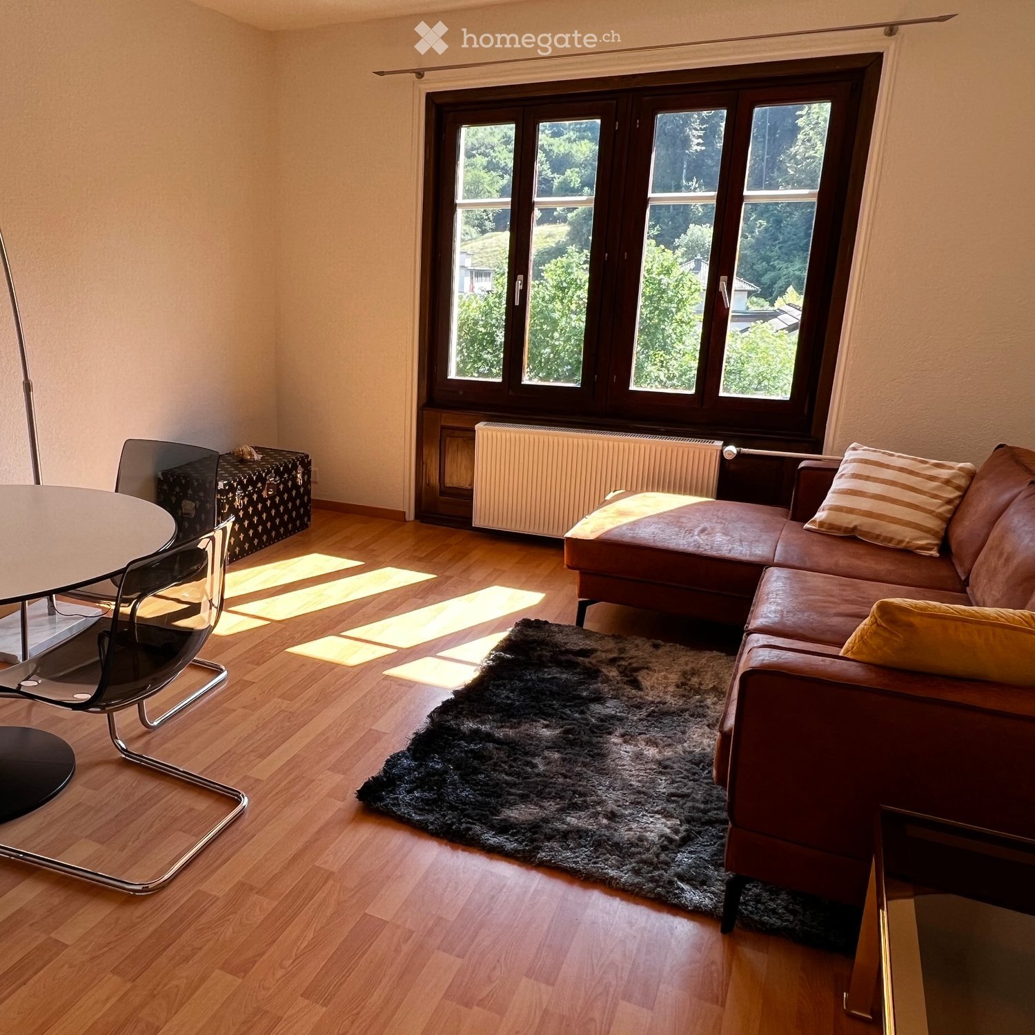 Living room with wooden flooring, large window, brown leather sofa, round glass table, modern chair, and a black shaggy rug.