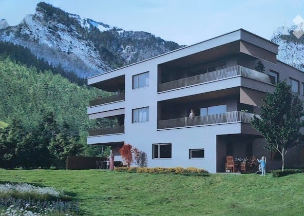 Modern multi-story building with balconies, surrounded by a well-maintained lawn and trees, with a scenic mountain view in the background.