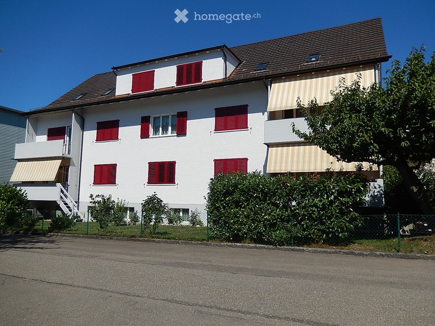 Two-story white house with red shutters, balconies, and striped awnings. The house has a tiled roof and is surrounded by greenery and a fence.